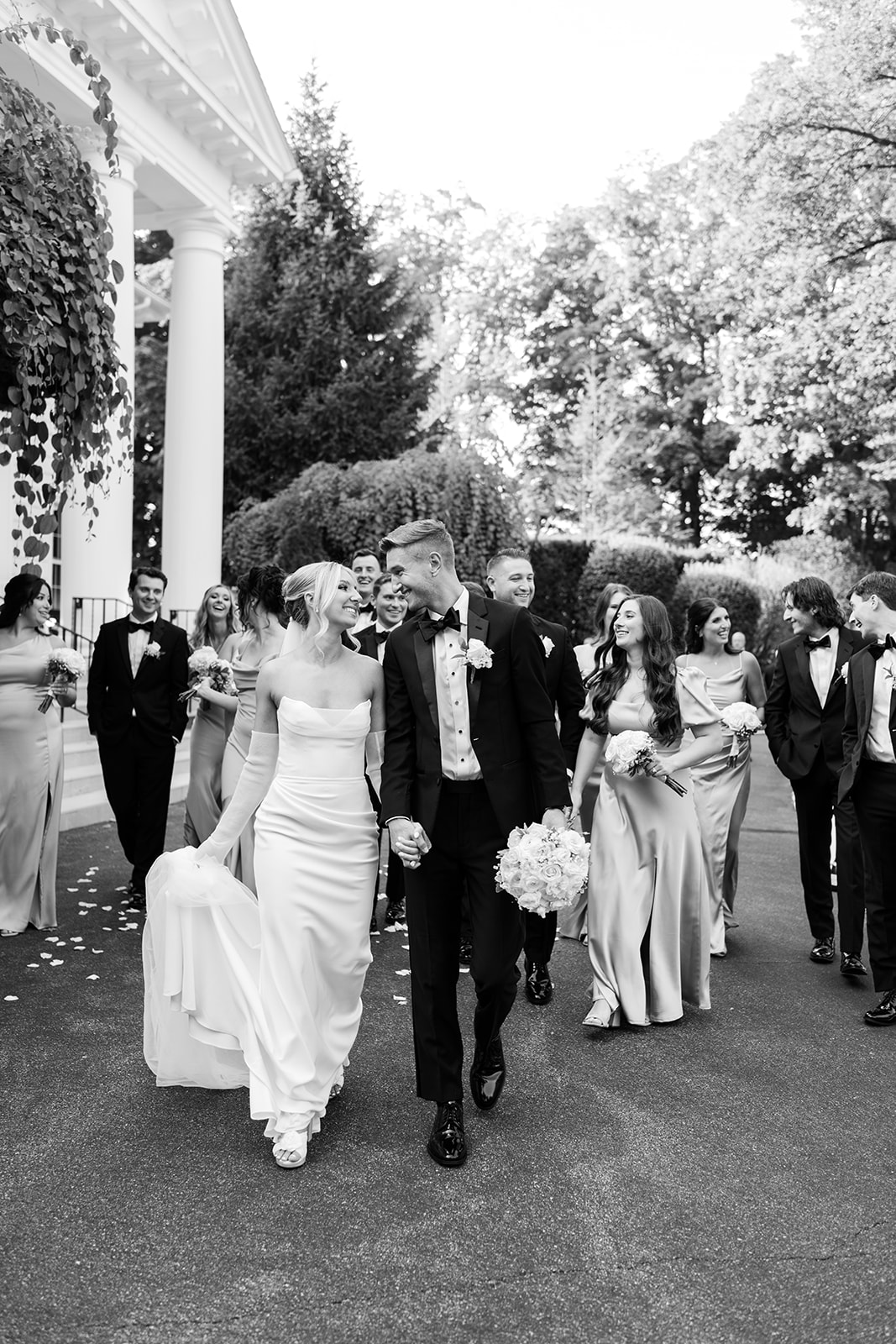 Black and white photo of the bride and groom walking hand-in-hand, leading their wedding party down a path.